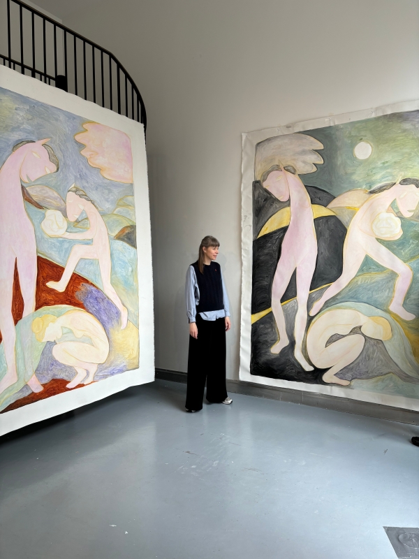 Artist
                                          Sarah Bogner stands in a high-ceilinged exhibition space between two large-scale paintings. The paintings depict stylized,
                                          pastel-colored figures with wing-like forms and, in some cases, horse-like heads, set within dreamlike landscapes. The artist
                                          is smiling. 